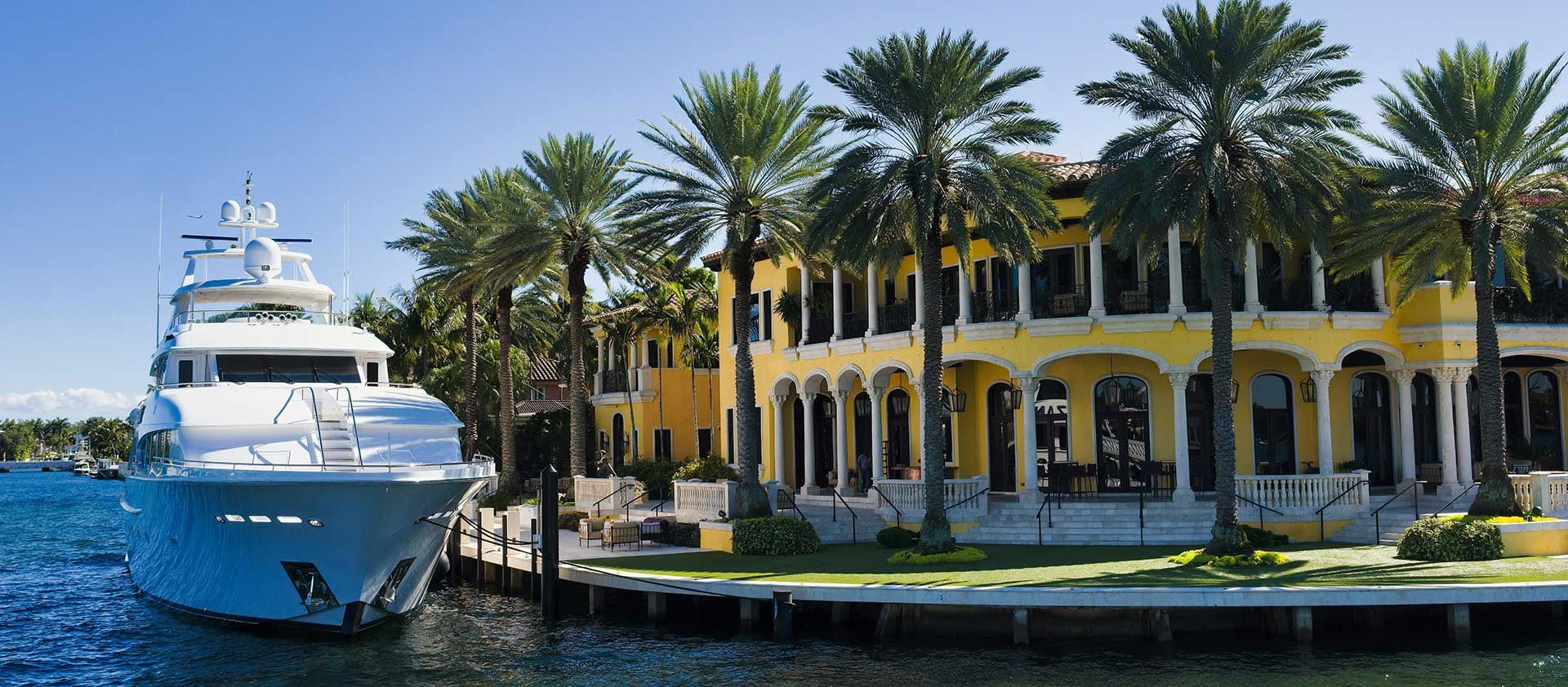 A white yacht is docked adjacent to a large, yellow mansion, separated by a small parkway adorned with full-grown palm trees and a well-manicured lawn.